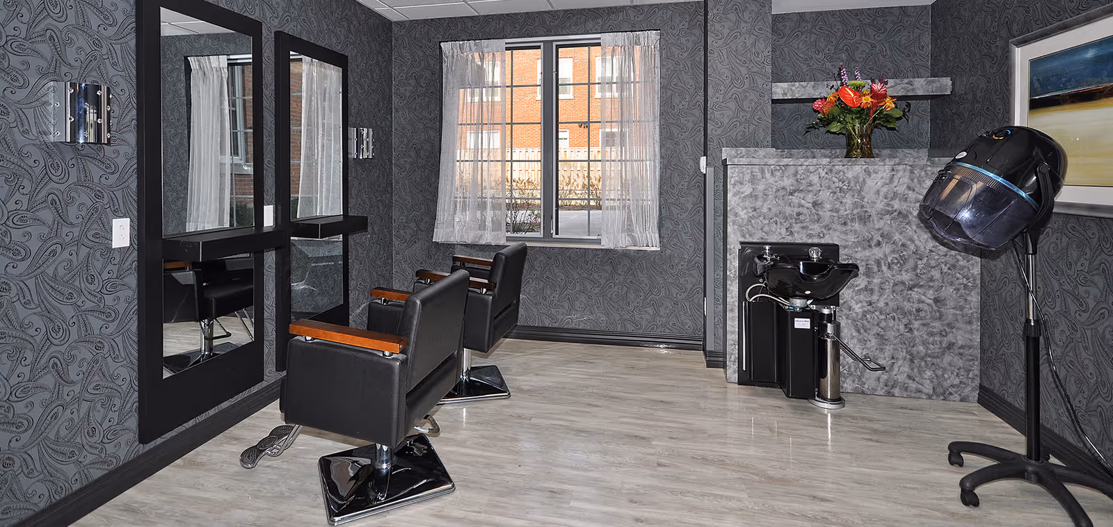 Interior view of a hair salon area with two black salon chairs facing large mirrors mounted on a patterned gray wall. There is a window with sheer white curtains letting in natural light. A hair washing station with a black sink and a hair dryer hood on a stand are visible. A vase with colorful flowers is placed on a gray textured counter.
