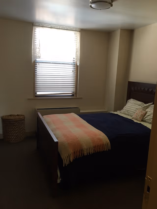 A small bedroom with a wooden bed frame, a bed made with a dark blue blanket, striped pillows, and a pink and cream checkered throw blanket. There is a window with blinds and lace curtains above a heating unit, and a woven basket is placed on the floor to the left of the bed.
