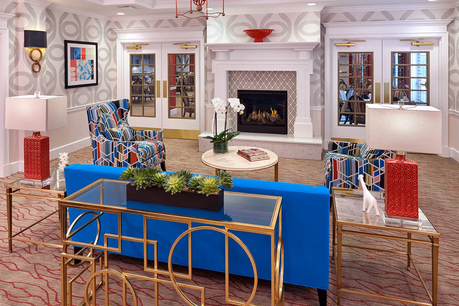 A cozy senior living facility lounge area featuring a blue sofa, two colorful patterned armchairs, a round coffee table with a white orchid plant and books, and a white fireplace with a fire burning. The room has patterned wallpaper, red lamps on side tables, and double glass doors leading to another room.