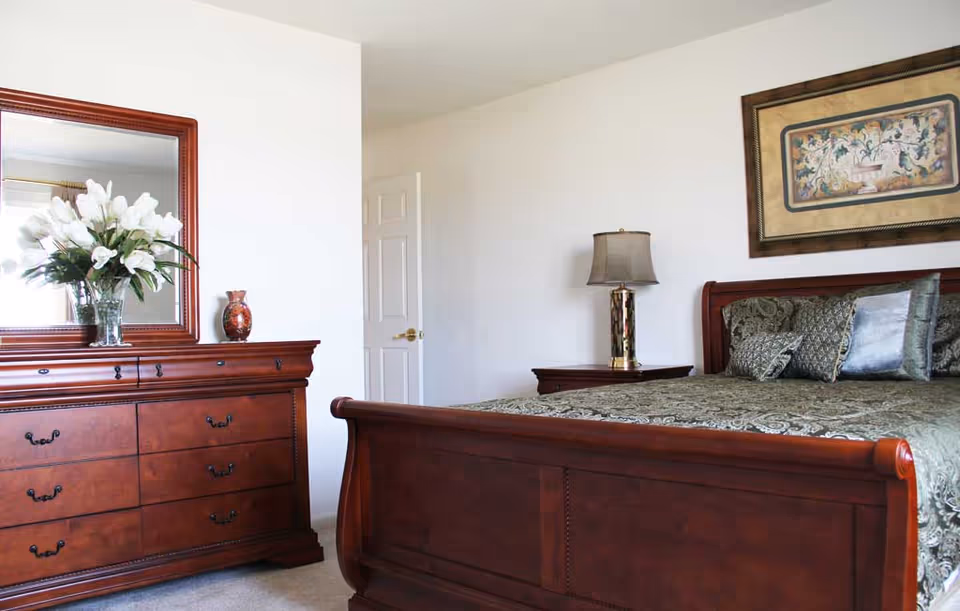 Bright bedroom featuring a wooden sleigh bed with patterned bedding, matching dresser and mirror, nightstand with lamp, and decorative accents.