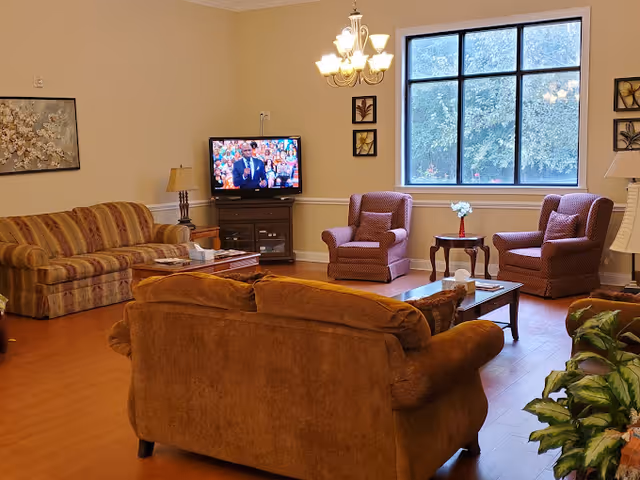 A cozy living room in a senior care facility with multiple upholstered chairs and sofas arranged around wooden coffee tables. A television is on, showing a man speaking, and there are framed artworks on the walls. A large window lets in natural light, and a chandelier hangs from the ceiling.
