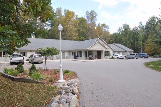 Single-story senior living building with a central entrance, parking area, lamp post, and trees in the background.