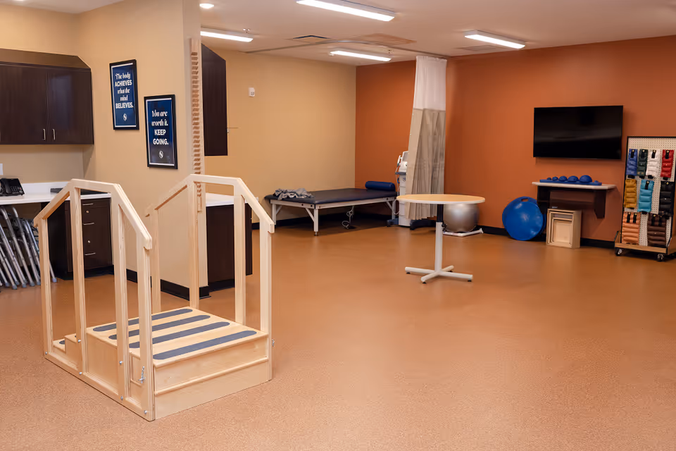 A spacious physical therapy room with wooden practice stairs, a treatment table, exercise equipment, and a wall-mounted TV.