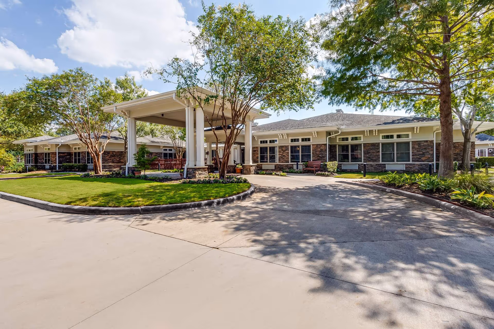 Front entrance of a single-story senior living building with a covered porte-cochere, landscaped lawn, and driveway.