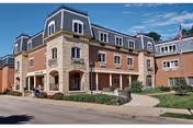 Front view of a multi-story brick and stone retirement center with a mansard roof, covered entrance, landscaping, and an American flag.