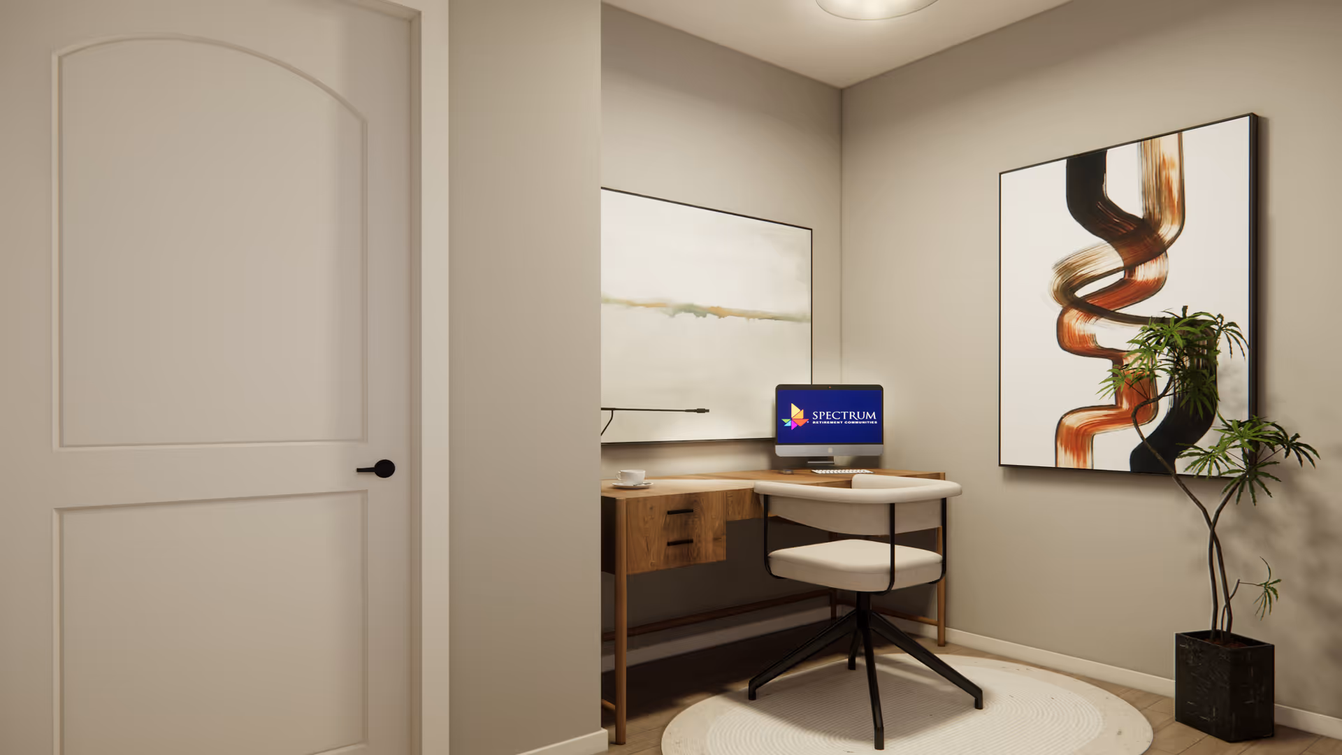 A small, modern home office corner with a wooden desk, a white cushioned swivel chair, a computer displaying the Spectrum Retirement Communities logo, two abstract paintings on the walls, and a potted plant in the corner.