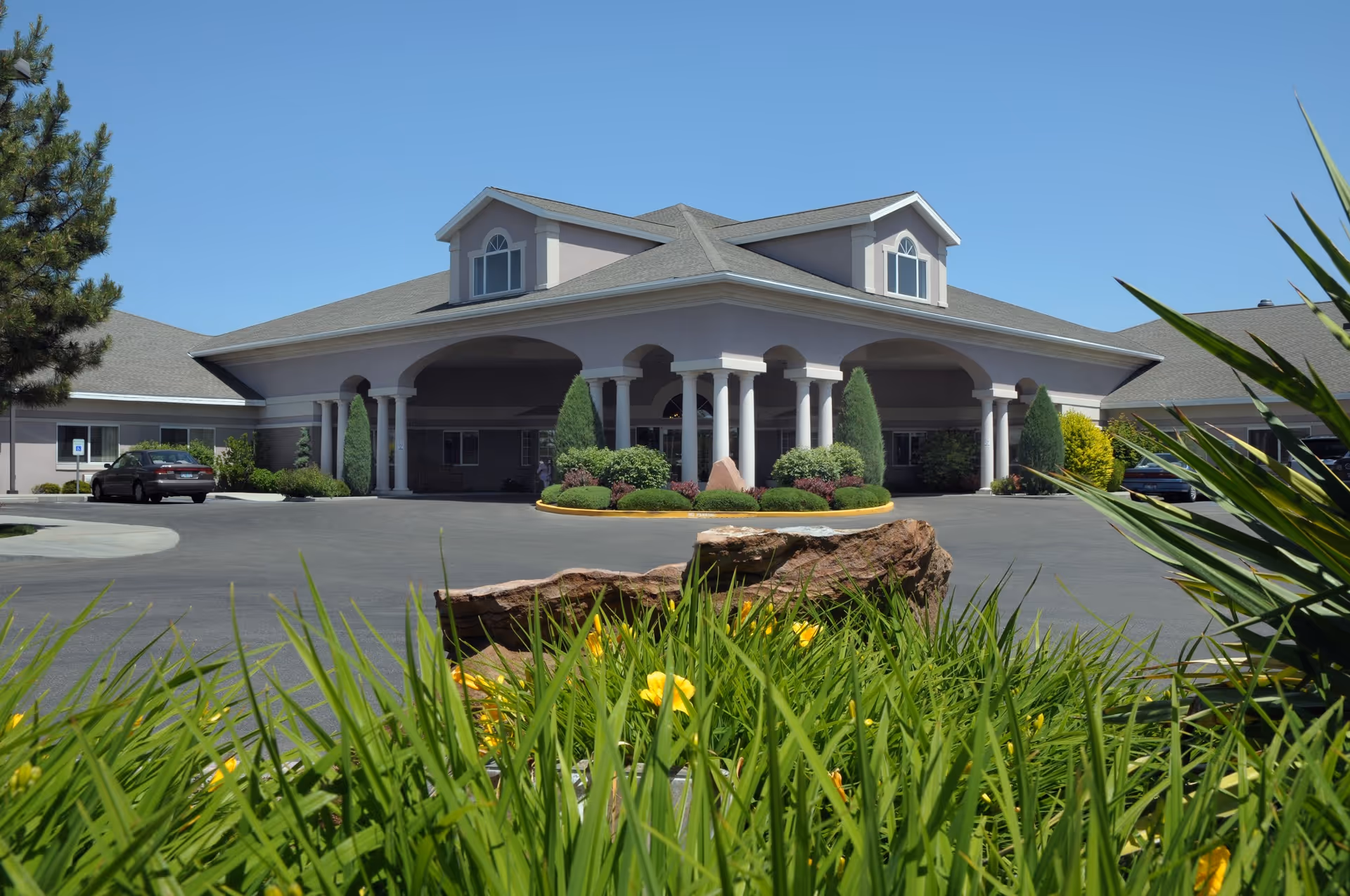 Front entrance of a senior living facility with a covered porte-cochère, columns, circular driveway, and landscaped foreground.
