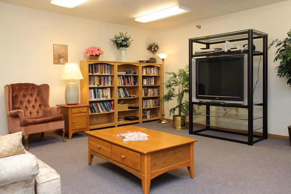 Cozy common room with bookshelves, armchairs, a wooden coffee table, and a television on a black stand.