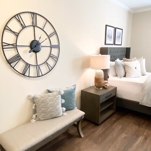 A cozy bedroom corner featuring a large decorative wall clock with Roman numerals, a cushioned bench with two pillows, a bedside table with a lamp and decorative items, and a neatly made bed with multiple pillows against a light-colored wall.