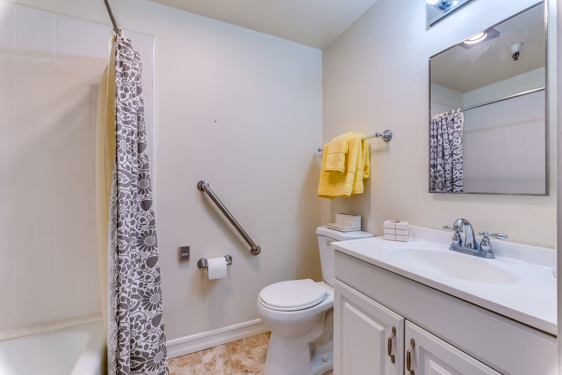 A clean bathroom featuring a white toilet with a yellow towel hanging above it, a white vanity with a sink and chrome faucet, a large mirror above the sink, a bathtub with a floral patterned shower curtain, and a grab bar on the wall next to the toilet.