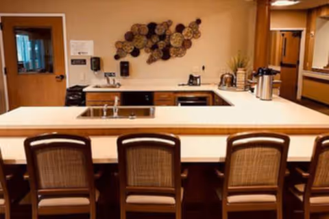 Communal kitchenette with a bar counter, four high-backed chairs, a sink, coffee urns and decorative wall art.