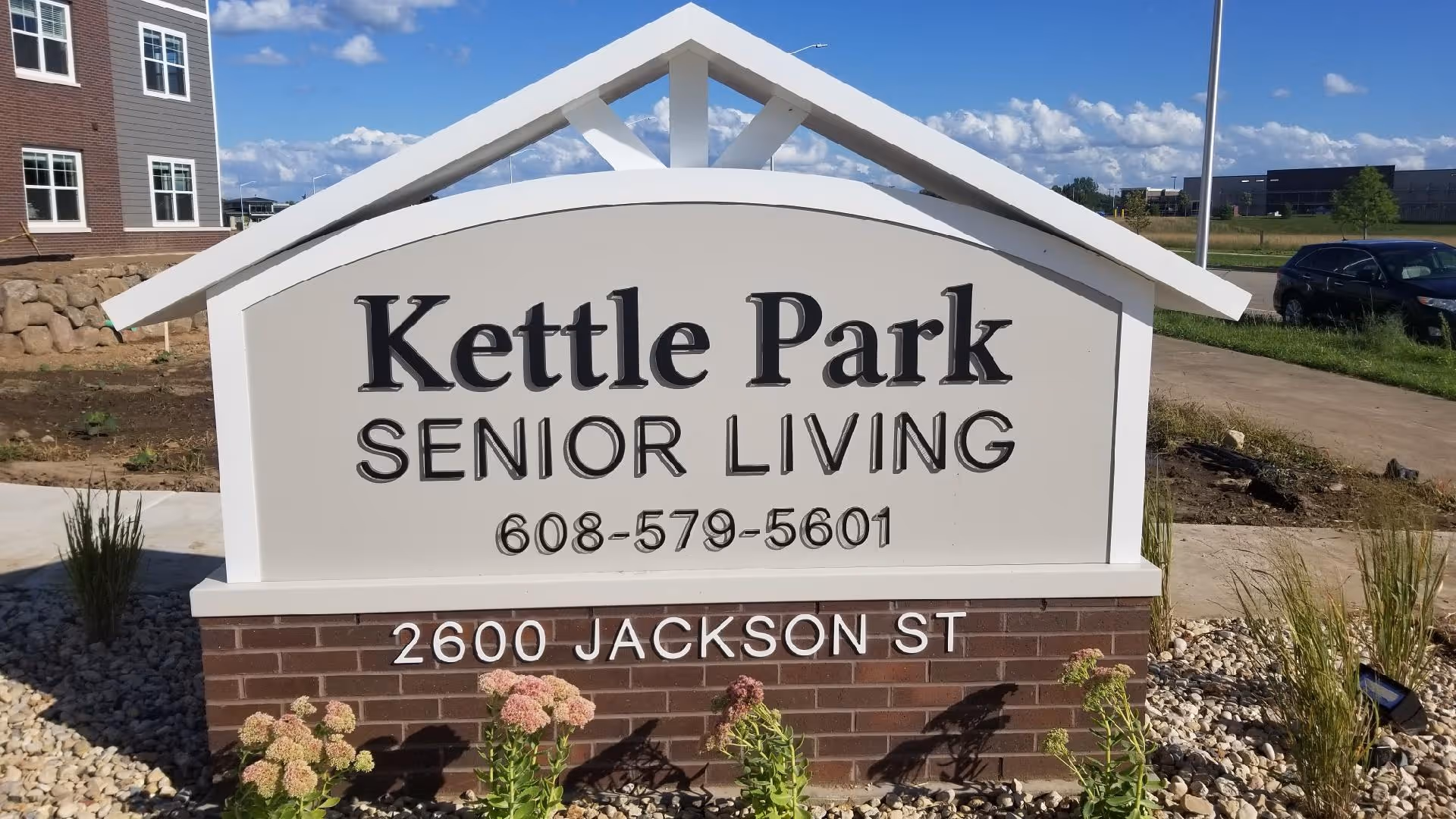 Freestanding entrance sign reading 'Kettle Park Senior Living 608-579-5601 2600 Jackson St' with a building and landscaping in the background.