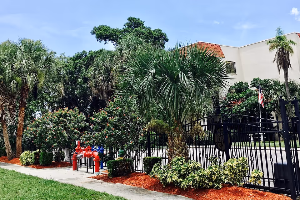 Exterior view of Langdon Hall facility showing a gated entrance with black metal fencing, surrounded by lush greenery including palm trees, bushes, and other plants. The building is light-colored with a red-tiled roof section and an American flag is visible near the entrance.