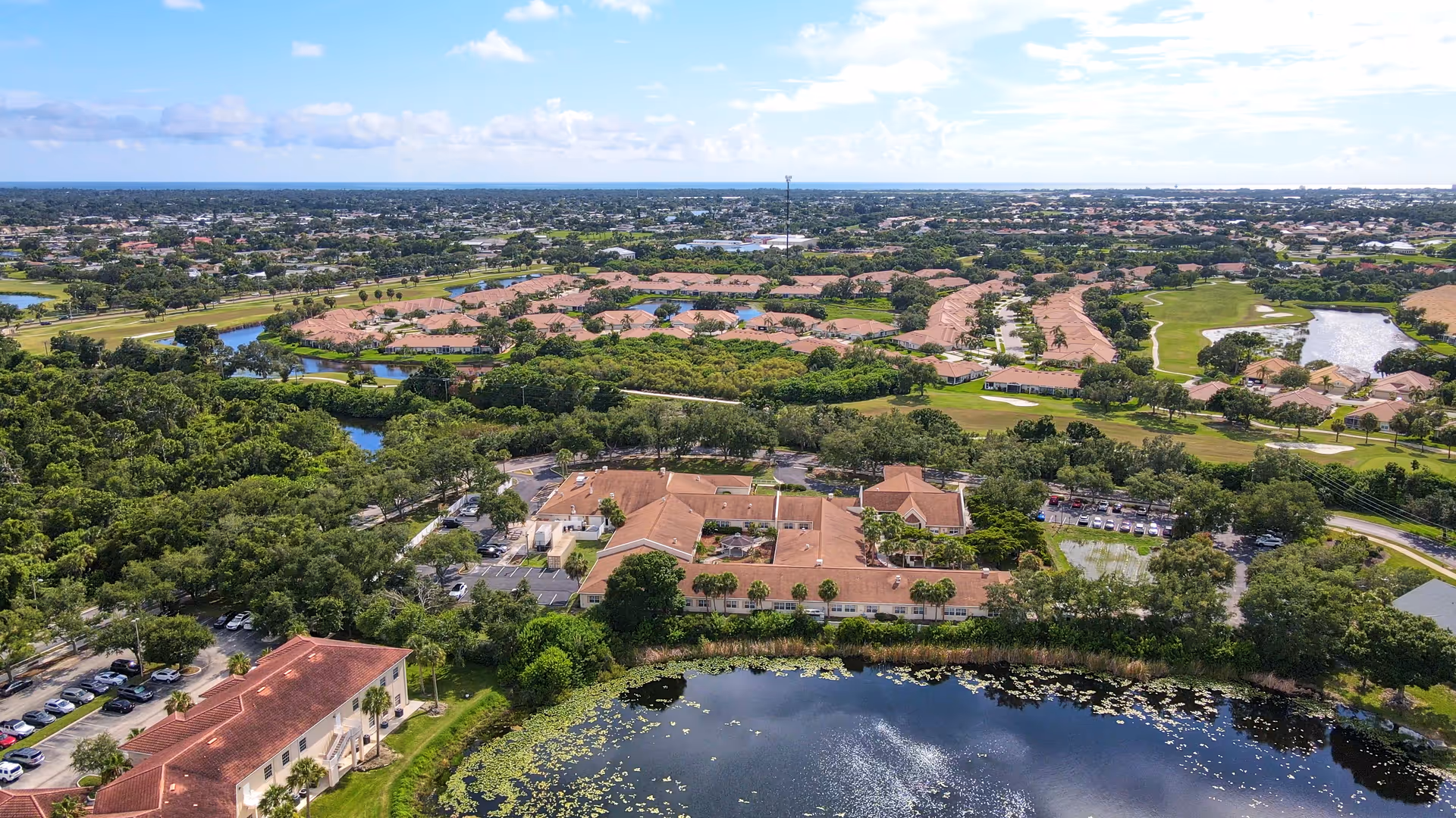Aerial view of Sunset Lake Health and Rehabilitation Center surrounded by trees, a pond with lily pads, and nearby residential areas with houses and green spaces under a partly cloudy sky.