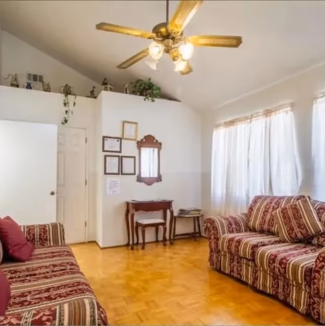 A cozy living room with two striped sofas facing each other, a small wooden table with a mirror above it on the wall, framed certificates or pictures, a ceiling fan with lights, and large windows with sheer curtains letting in natural light.