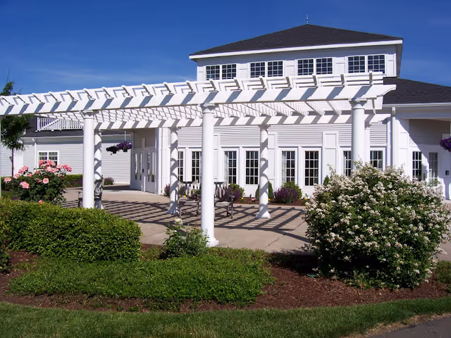 Outdoor patio area with white pergola structure casting shadows on the ground, surrounded by green bushes and flowering plants, in front of a white building with multiple windows under a clear blue sky.