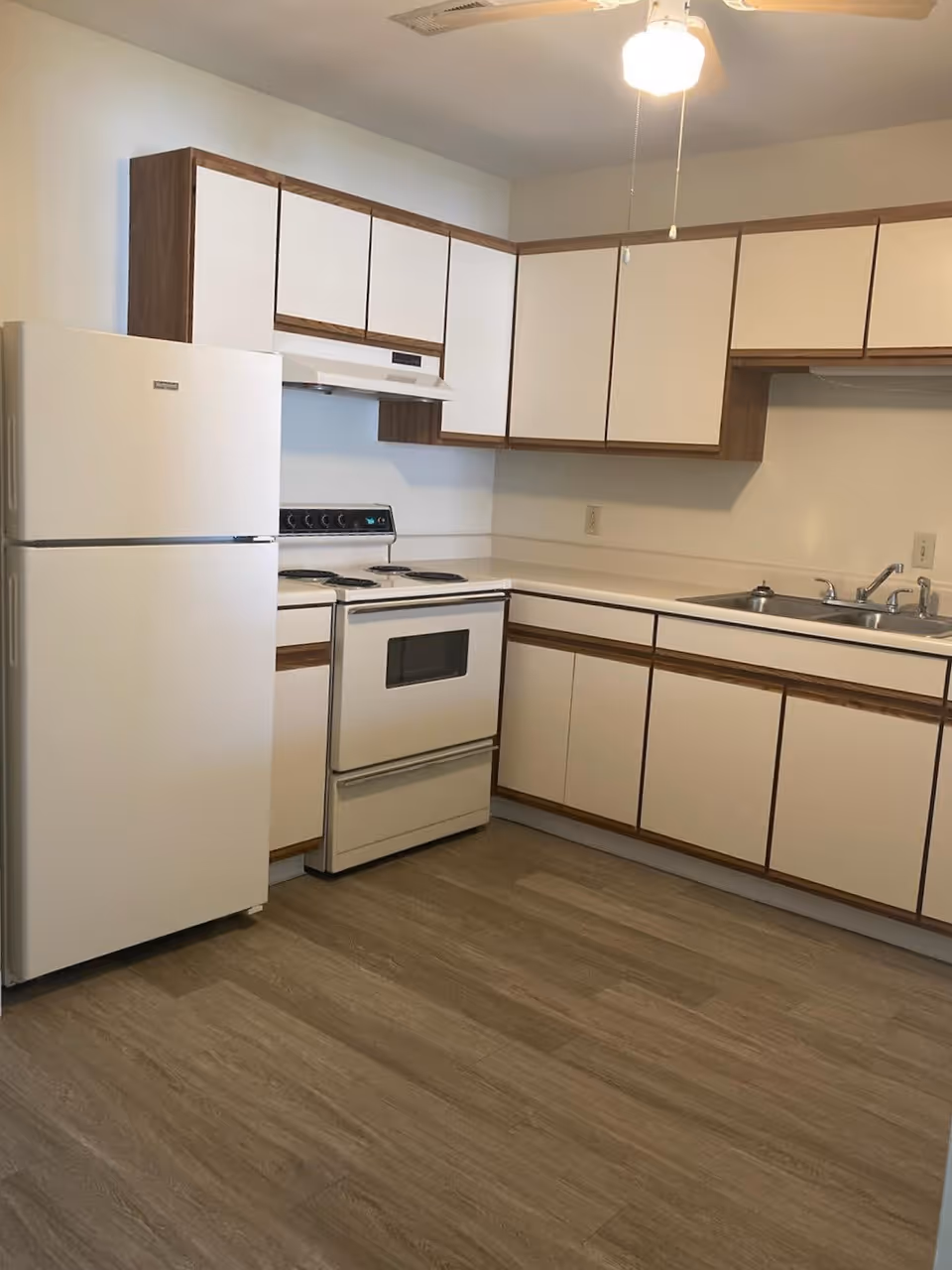 A kitchen with white cabinets featuring wood trim, a white refrigerator, an electric stove with oven, a double sink, and a ceiling fan with a light. The floor has a wood-like finish.