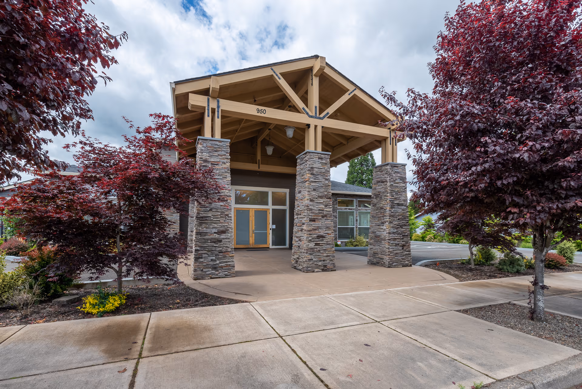 Entrance of a building with a covered porch supported by four stone pillars. The building has a wooden roof structure and glass doors. There are red-leaved trees and landscaping on either side of the entrance, with a concrete sidewalk and driveway in front. The number 950 is visible on the wooden beam above the entrance.