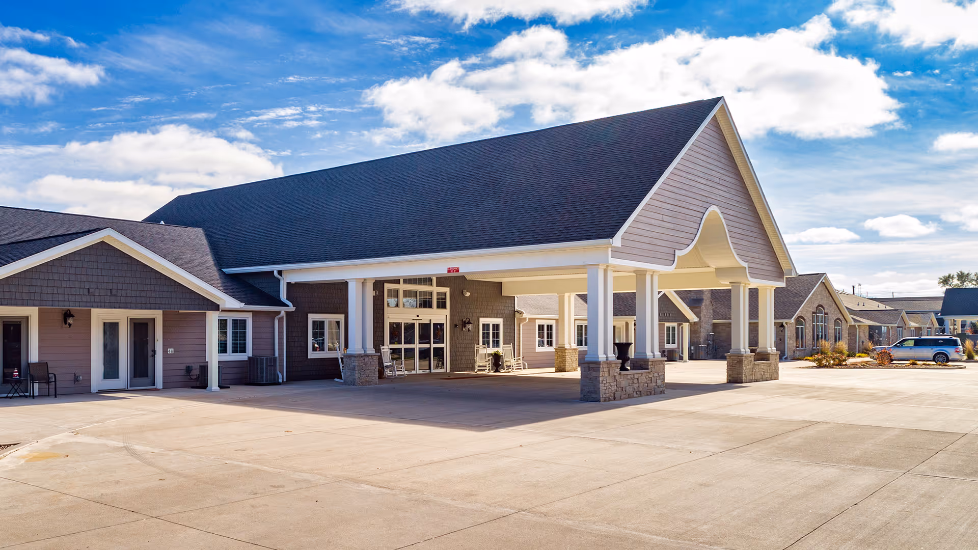 Exterior view of Villas of Holly Brook Assisted Living & Memory Care in Pekin, IL, showing a large covered entrance with white columns and stone bases, multiple windows, and a clear blue sky with some clouds.