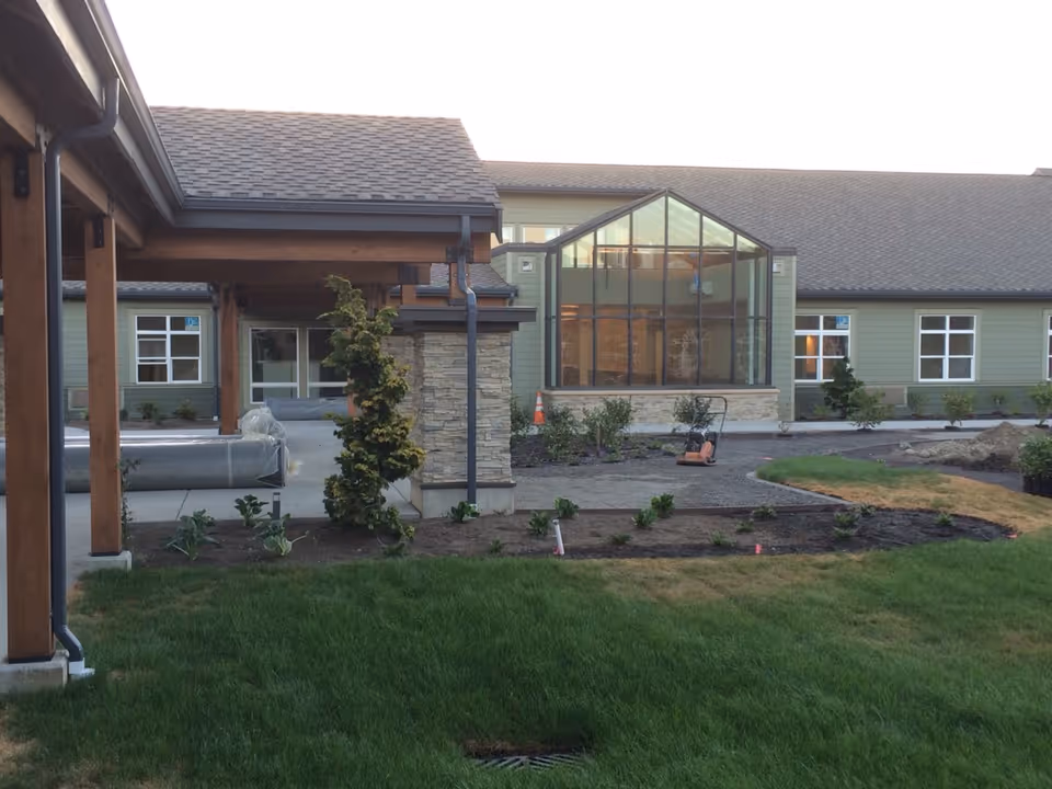 Exterior courtyard and entrance of a single-story memory care building with a covered wooden walkway, glass sunroom, and landscaped lawn.