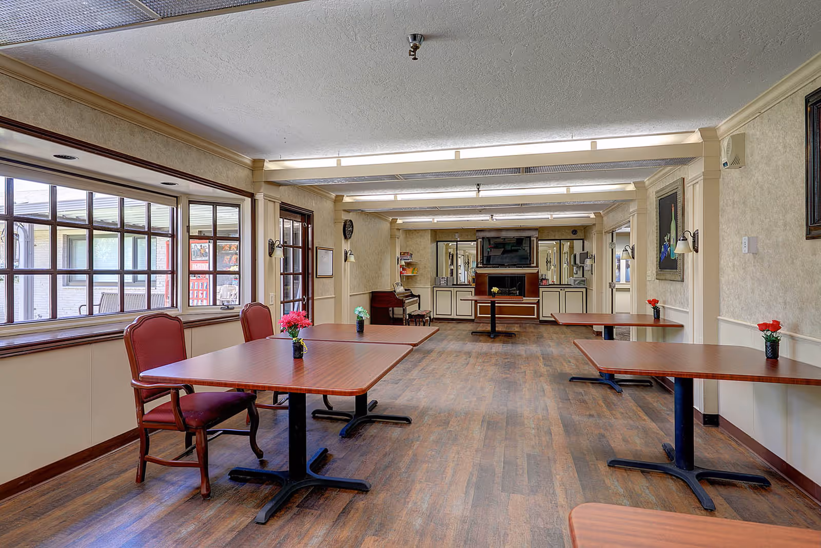Interior view of a dining room in a senior living facility with wooden tables and red cushioned chairs. Small flower vases are placed on the tables. Large windows on the left side allow natural light to enter the room. The room has wood flooring and beige walls with decorative molding. A television and additional seating are visible at the far end of the room.