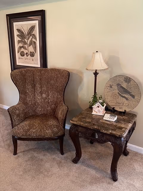 A cozy corner featuring a patterned upholstered wingback chair next to a dark wooden side table with a marble top. On the table are a small table lamp with a white shade, a decorative bird-themed round plaque, a small house-shaped ornament, a plant, and a remote control. A framed botanical print hangs on the beige wall behind the chair.