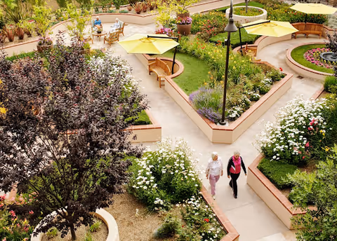 A landscaped outdoor garden area with paved walkways, raised flower beds, benches, and yellow umbrellas. Two elderly women are walking along the path, and two other people are seated at a table in the background. The garden is lush with various plants and flowers.