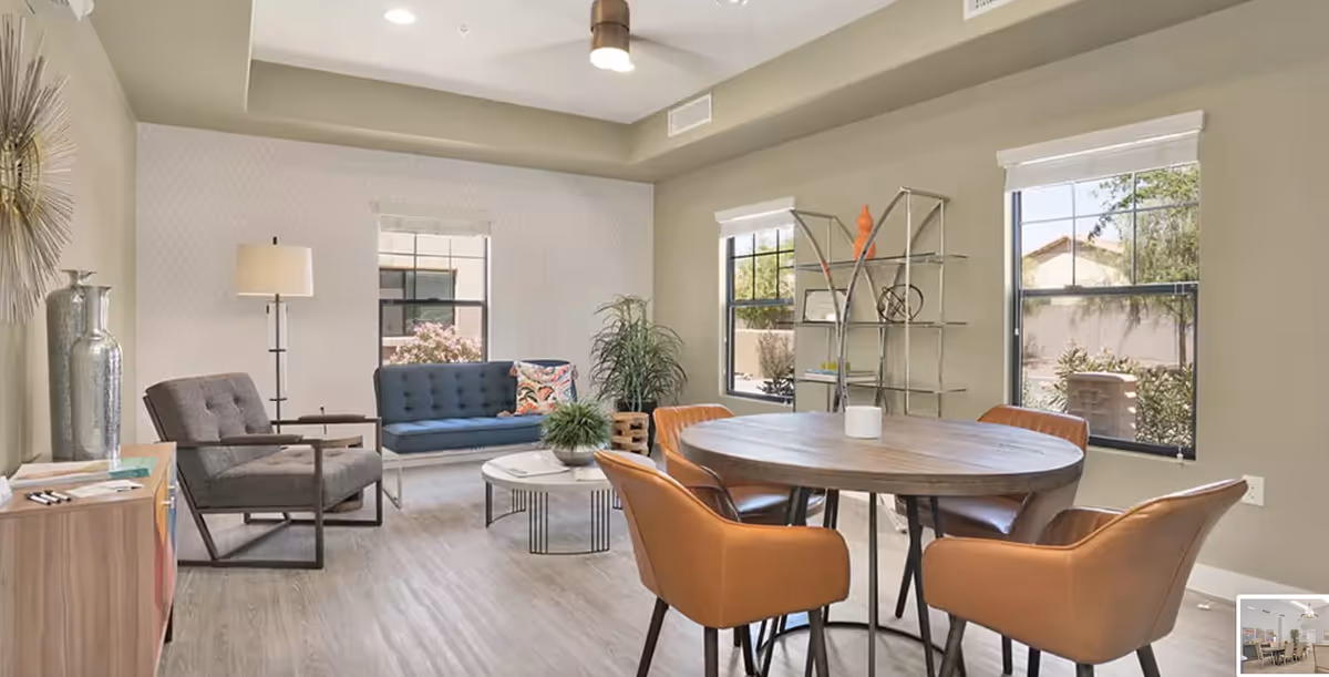 A bright and modern living room area with a round wooden table surrounded by four brown leather chairs. In the background, there is a blue cushioned bench, a gray armchair, a white round coffee table with a small plant, a tall floor lamp, and a metal shelving unit with decorative items. The room has large windows letting in natural light and light-colored walls and flooring.