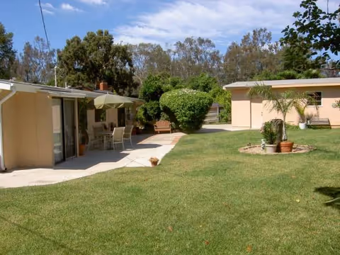 Outdoor lawn with a patio, chairs, umbrella, and beige villa-style buildings.