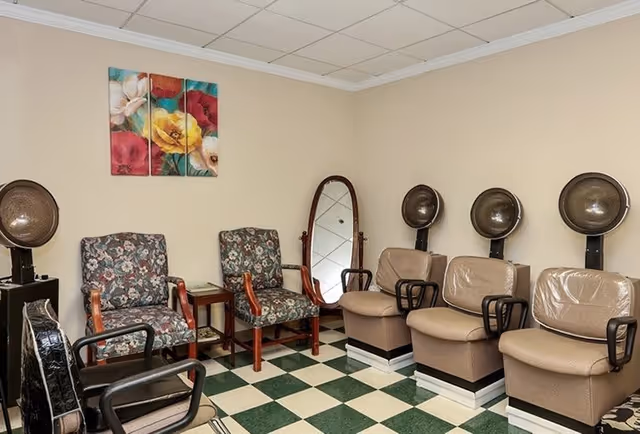 Interior of a hair salon area with three beige salon chairs equipped with hair dryers, two floral-patterned armchairs, a standing mirror, and a colorful floral painting on the wall. The floor has a green and white checkered pattern.