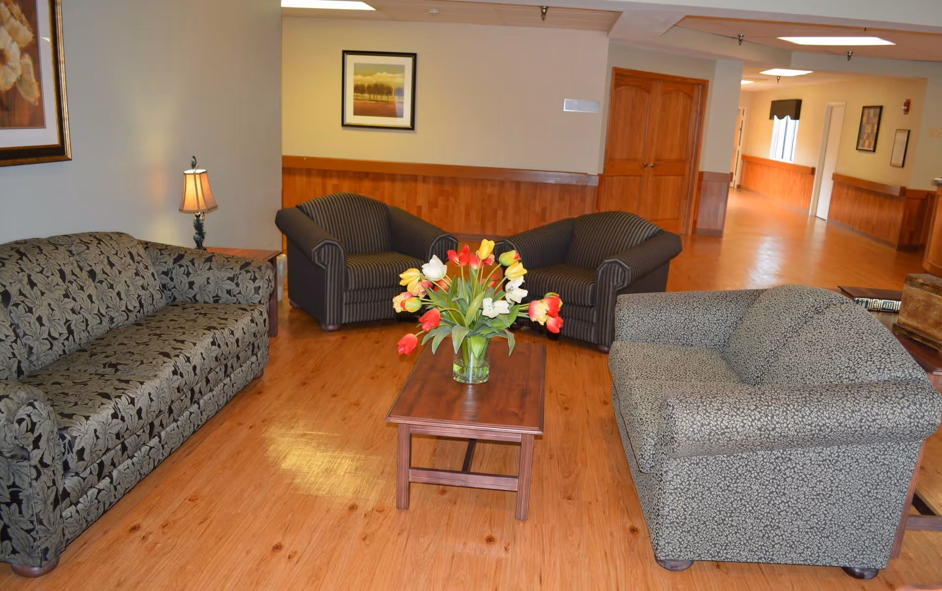 A cozy sitting area in a nursing home with three upholstered sofas arranged around a wooden coffee table holding a vase of colorful tulips. The room has wooden flooring, light-colored walls with wood paneling, a small table lamp, framed artwork on the walls, and a hallway leading to other rooms.