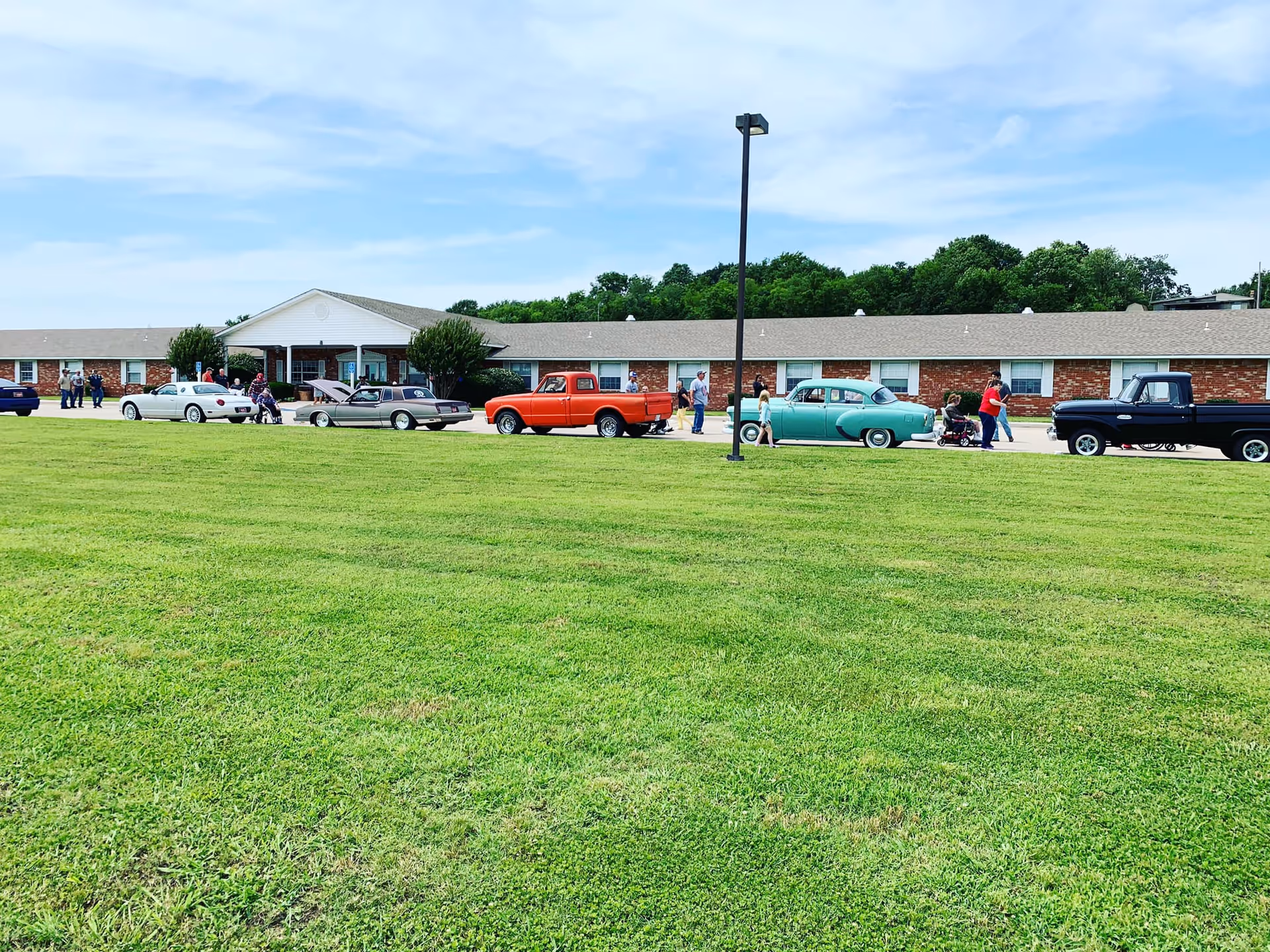 A single-story brick building with a white entrance canopy, several vintage cars and trucks parked in front, and people walking and standing near the vehicles on a sunny day with a blue sky and some clouds. There is a large green lawn in the foreground and a tall street lamp near the center.