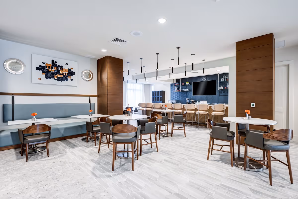 Modern dining area with round and rectangular tables, wooden chairs with gray cushions, and a long bench with blue upholstery. The room features two large wooden columns, pendant lights hanging from the ceiling, a bar area with stools, a flat-screen TV mounted on a dark blue textured wall, and decorative wall art.