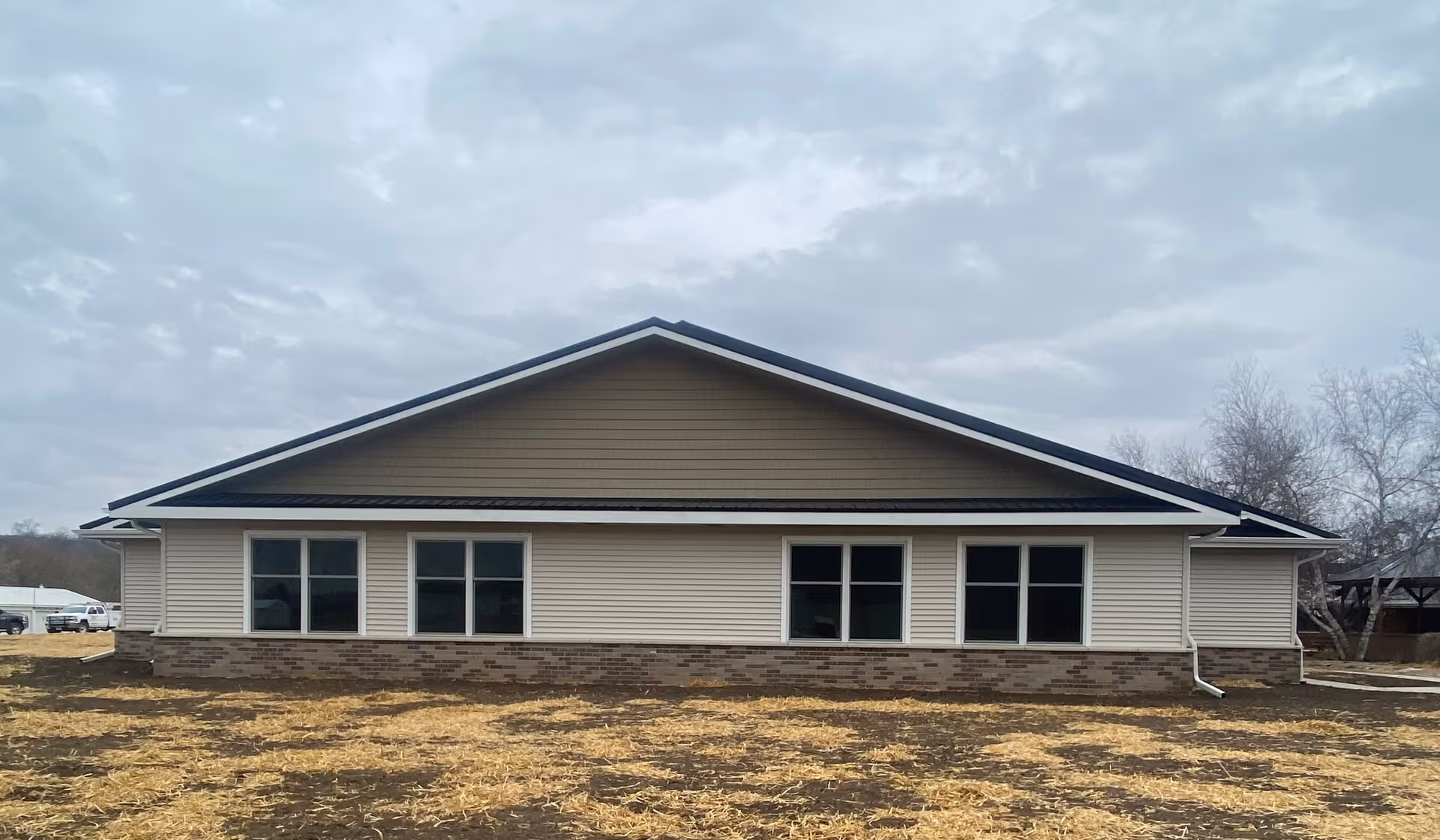 Exterior view of a single-story building with beige siding, a brown brick base, and a dark brown triangular roof under a cloudy sky. The ground in front is covered with straw and patches of dirt, and there are leafless trees visible on the right side.