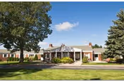 Exterior view of a single-story brick building with a covered entrance, surrounded by green grass, trees, and a clear blue sky with a few clouds.