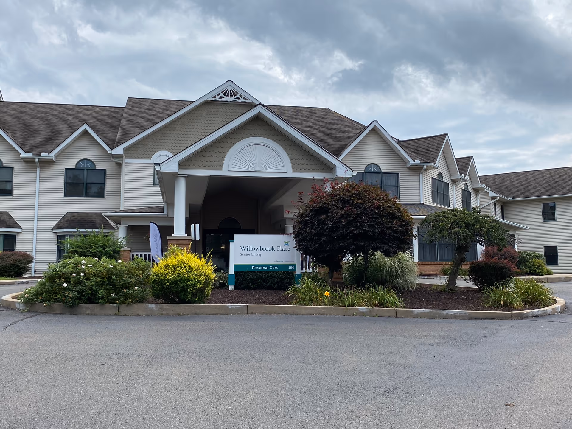 Exterior front view of Willowbrook Place senior living facility with a covered entrance, landscaped bushes and trees, and a sign indicating personal care services.