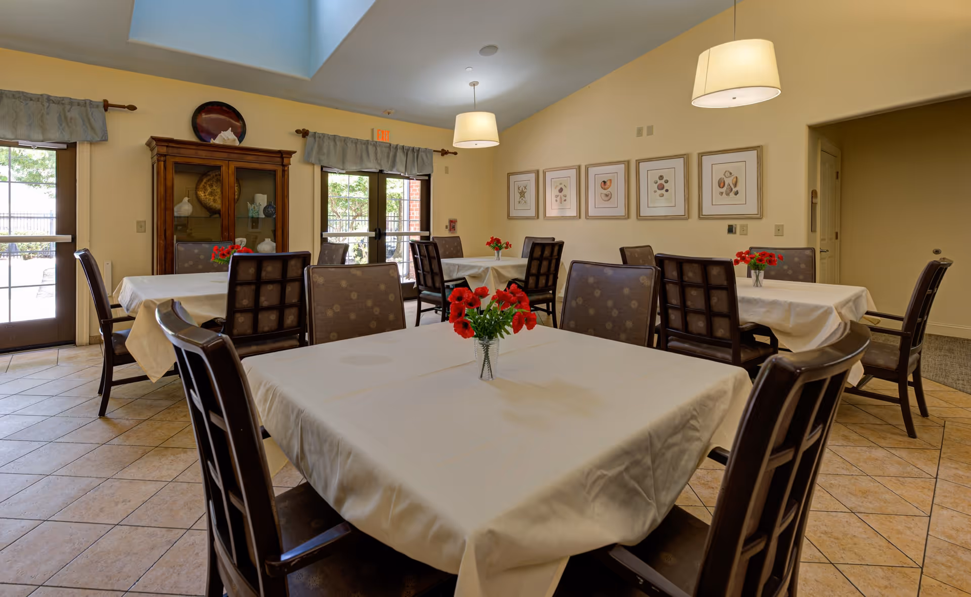 A dining room with several tables covered in white tablecloths, each adorned with a small vase of red flowers. The room has tiled floors, beige walls, framed artwork on the wall, and large windows and doors letting in natural light. There are cushioned chairs around the tables and two pendant lights hanging from the ceiling.