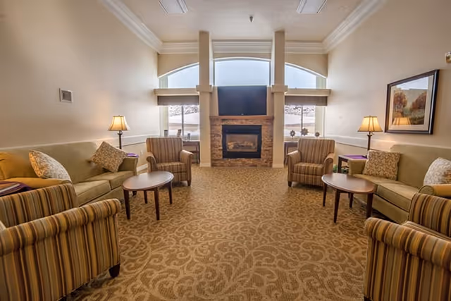 A cozy living room in an assisted living facility with two beige sofas and four striped armchairs arranged around two small wooden coffee tables. The room features a carpet with a subtle pattern, a stone fireplace with a flat-screen TV mounted above it, two table lamps on side tables, and large windows letting in natural light.
