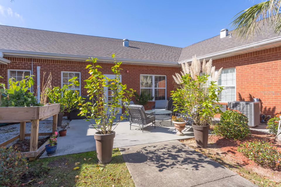 Outdoor patio area at Truewood by Merrill, Ocean Springs with potted plants, outdoor seating including chairs and a table, surrounded by a red brick building under a clear blue sky.