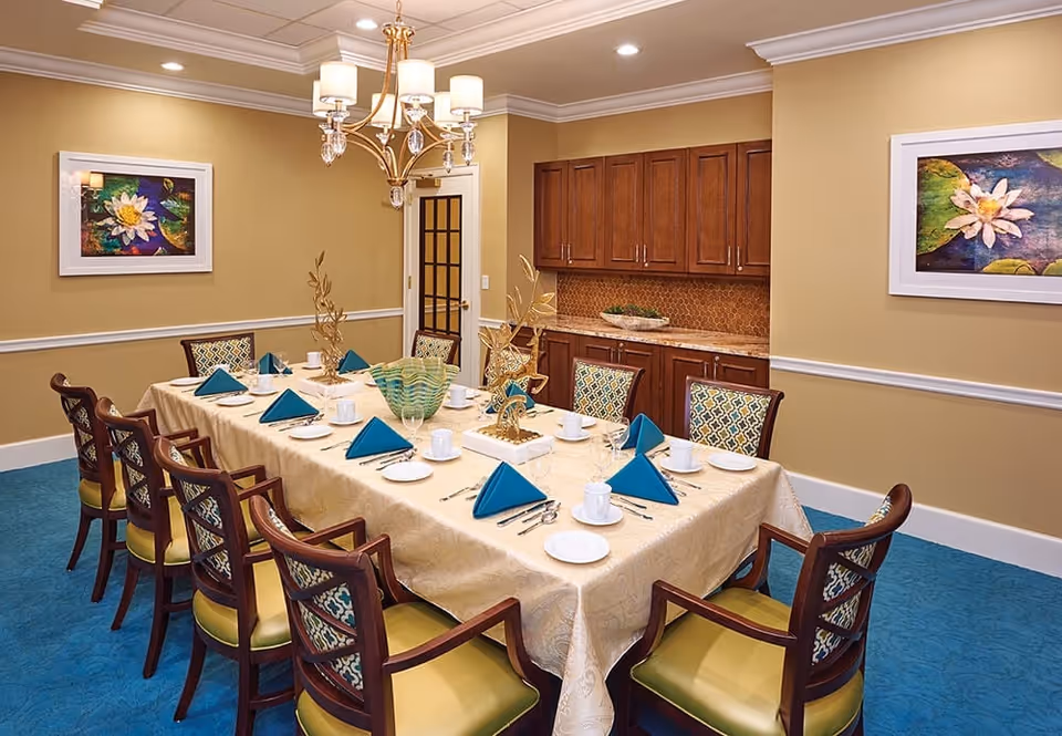 A formal dining room with a long rectangular table covered with a beige tablecloth, set with white plates, cups, silverware, and teal folded napkins. The room has wooden chairs with patterned upholstery, a blue carpet, beige walls with white trim, two framed floral paintings, wooden cabinets with a countertop, and a chandelier hanging from the ceiling.