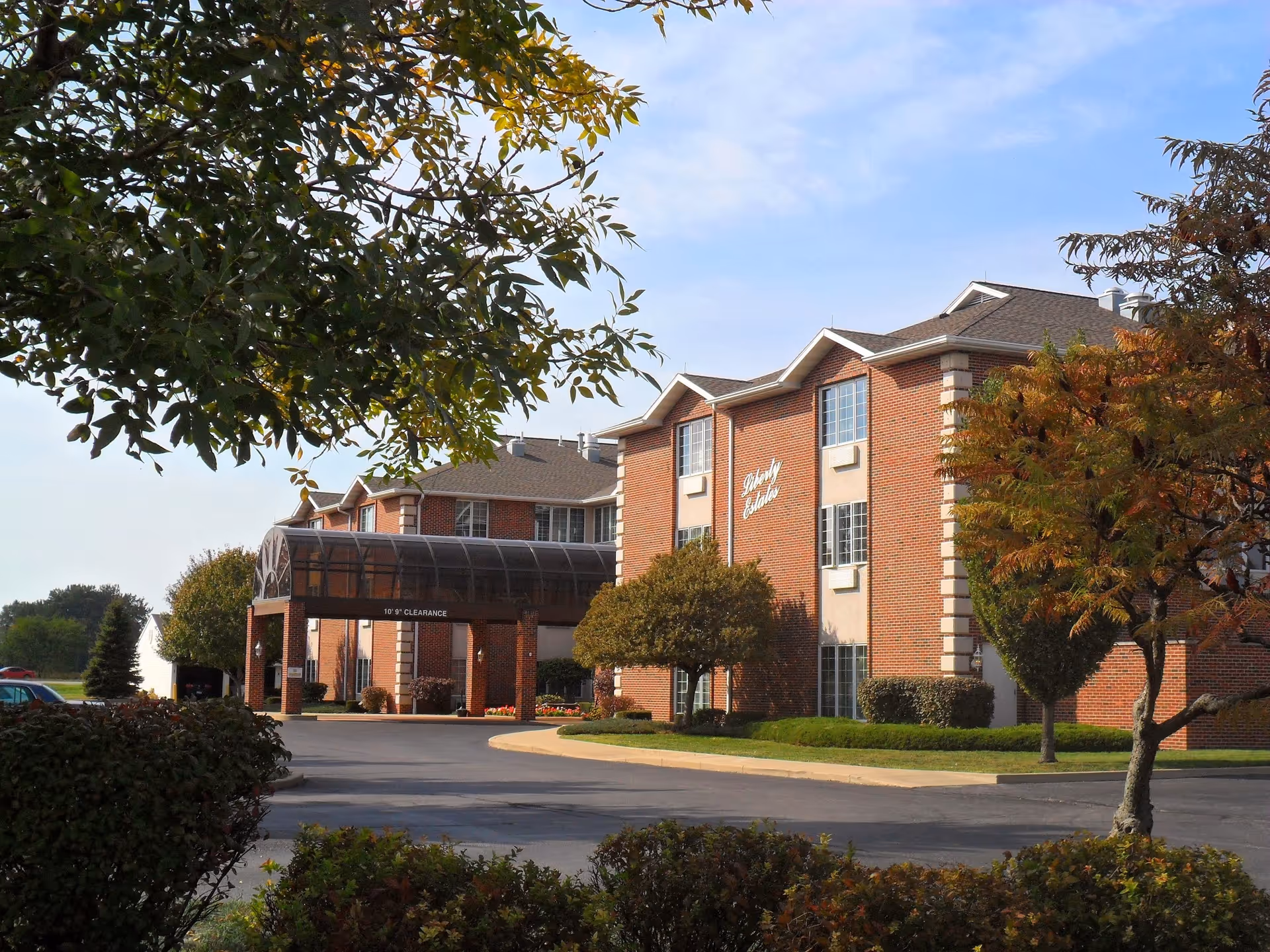 Exterior view of a three-story brick building with a covered entrance driveway. The building is surrounded by trees and shrubs, with a clear sky above. The sign on the building reads 'Liberty Estates'.