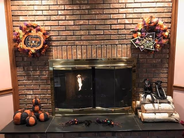 A brick fireplace with a glass and brass screen. On the mantel are two Halloween-themed wreaths, one with a 'Trick or Treat' sign and the other with a 'This Way to Treats' sign. To the left of the fireplace are decorative pumpkins in black and orange. To the right are two skeleton figurines sitting on a stack of white birch logs. A small Halloween garland is laid out in front of the fireplace.