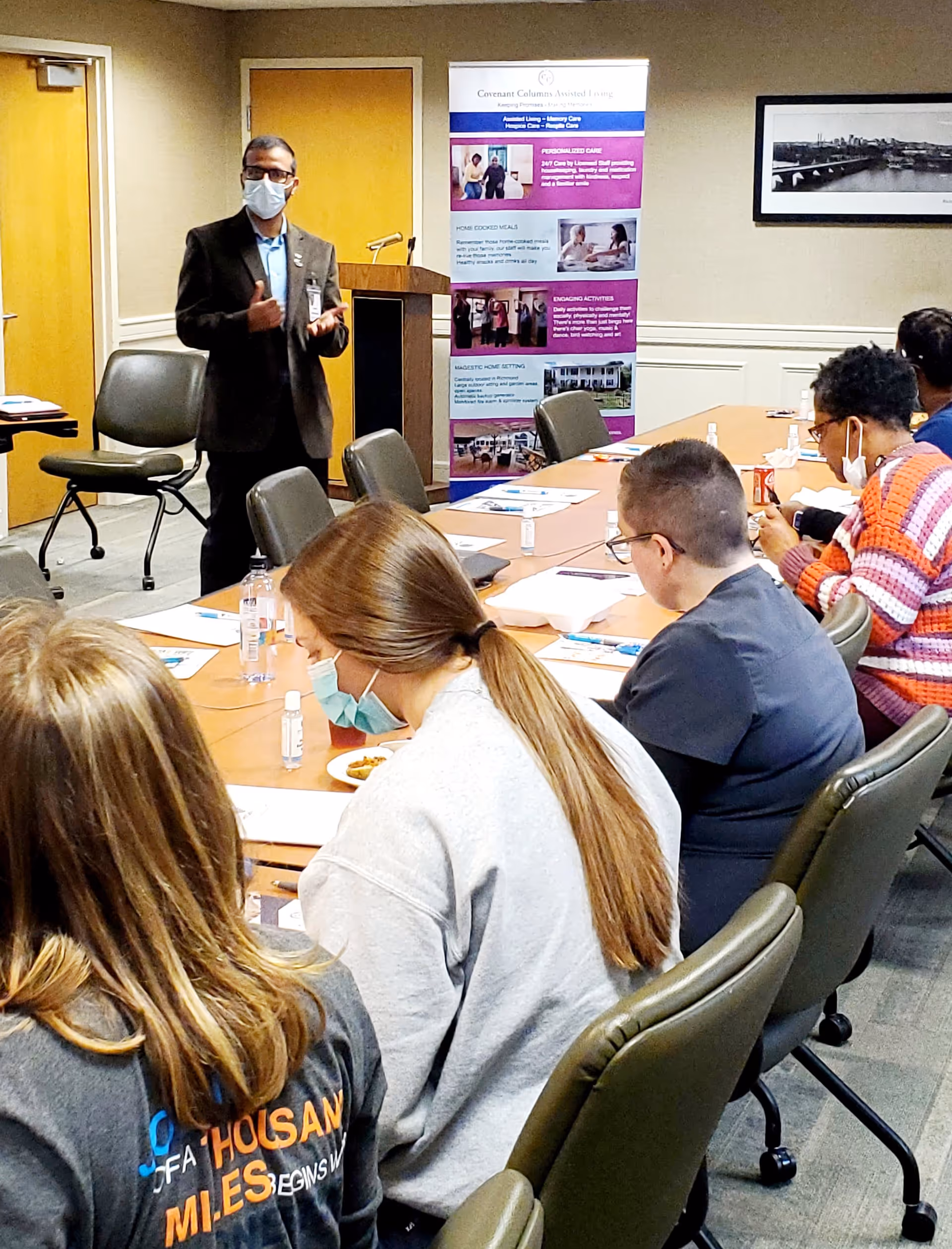 A man wearing a mask and suit stands at the front of a conference room giving a presentation to a group of seated people. The attendees, some wearing masks, are seated around a long table with papers, hand sanitizer, and drinks. Behind the presenter is a banner with information about Covenant Columns Assisted Living.