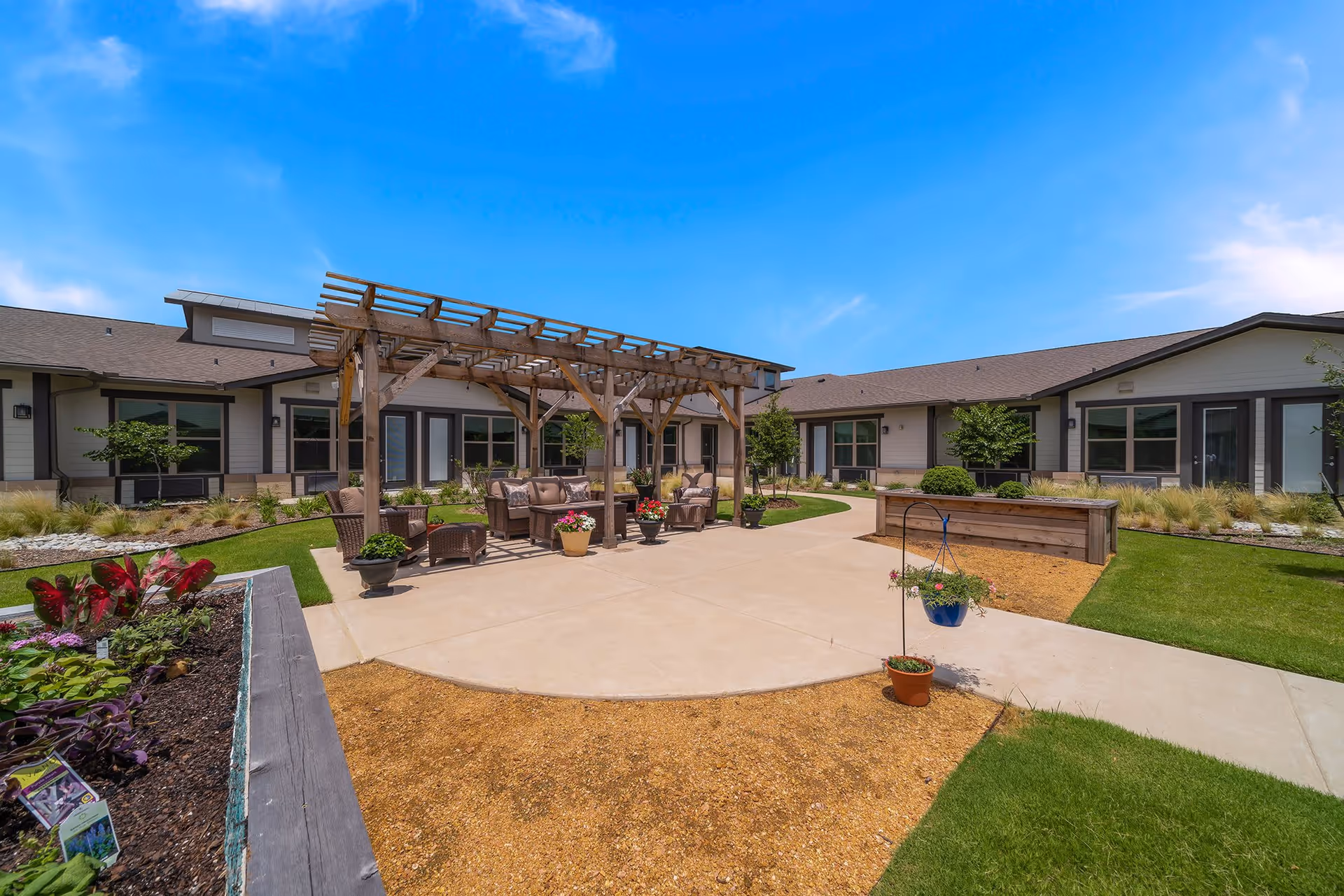 Outdoor courtyard area at Hidden Springs of McKinney featuring a wooden pergola with cushioned seating underneath, surrounded by landscaped garden beds, potted plants, and a paved walkway. The courtyard is enclosed by single-story buildings with large windows under a clear blue sky.