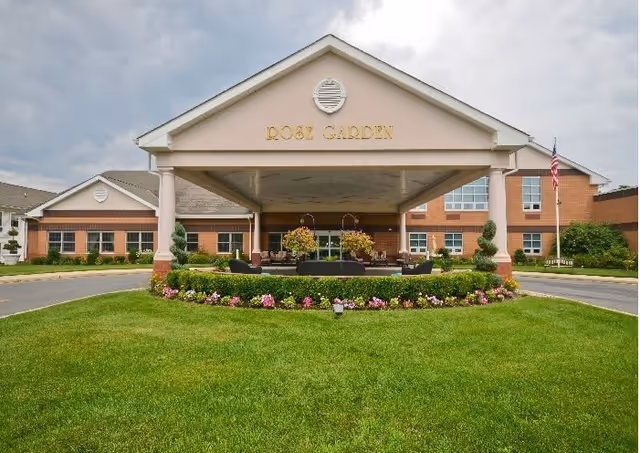 Front exterior view of Rose Garden Nursing and Rehabilitation Center showing a covered entrance with columns, a circular driveway, well-maintained green lawn, flower beds, and an American flag on a flagpole to the right.