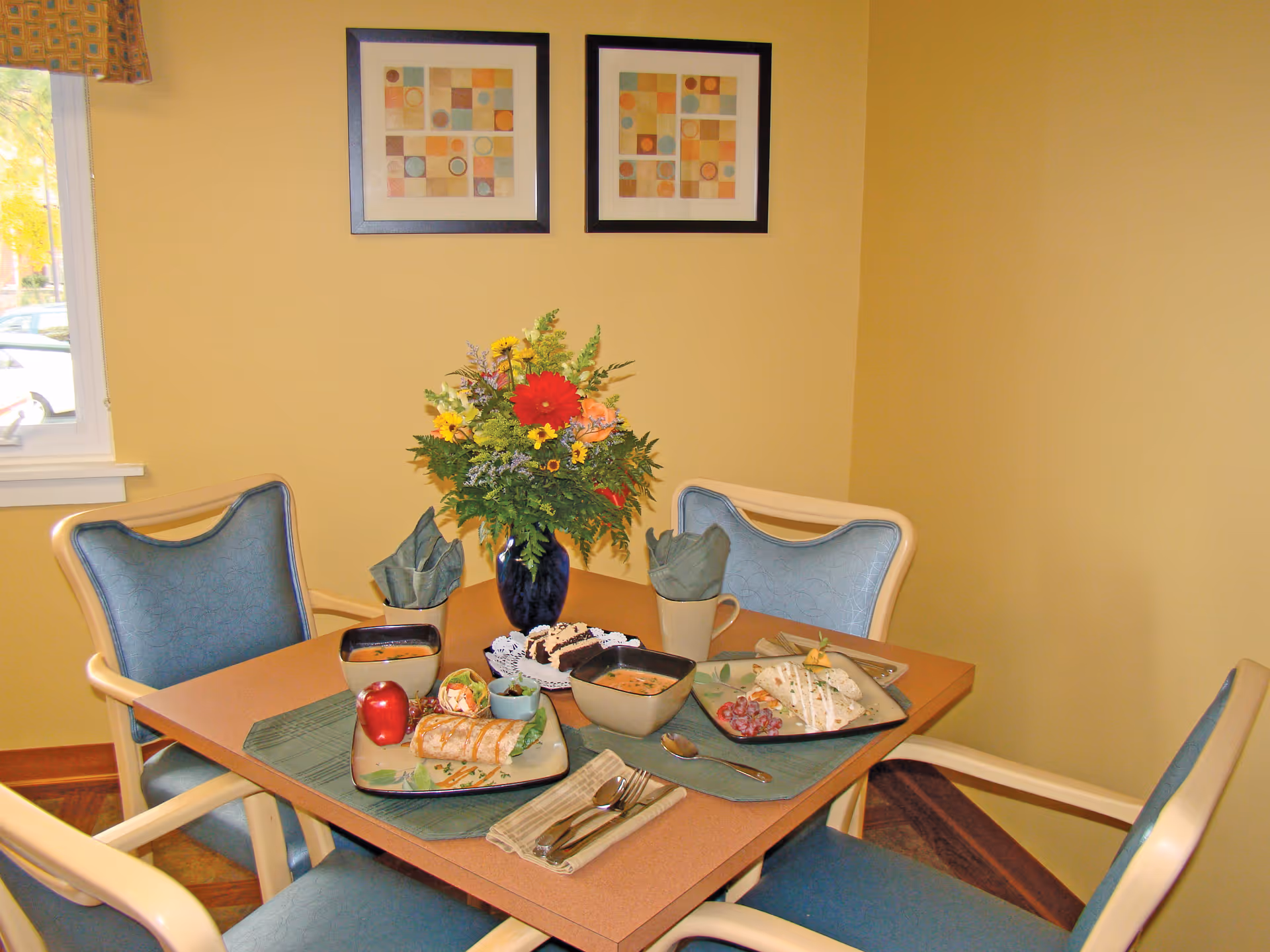 A small dining area with a square table set for two, featuring plates with wraps, bowls of soup, a red apple, and a dessert plate with brownies. The table has a green placemat and napkins, and a vase with a colorful flower arrangement is in the center. Four blue cushioned chairs surround the table. The walls are painted yellow, and two framed abstract art pieces hang on the wall. A window with a patterned valance is visible on the left side.