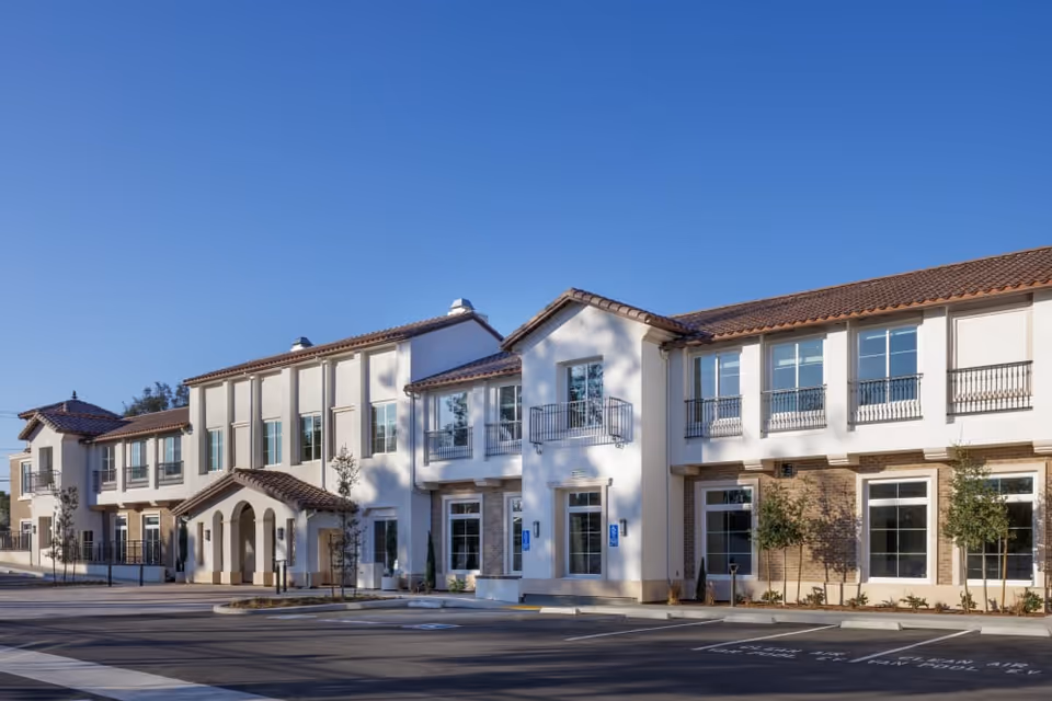 Exterior view of a two-story senior living facility building with white walls, large windows, and a tiled roof under a clear blue sky. The building has a covered entrance and several small trees planted around the parking area.