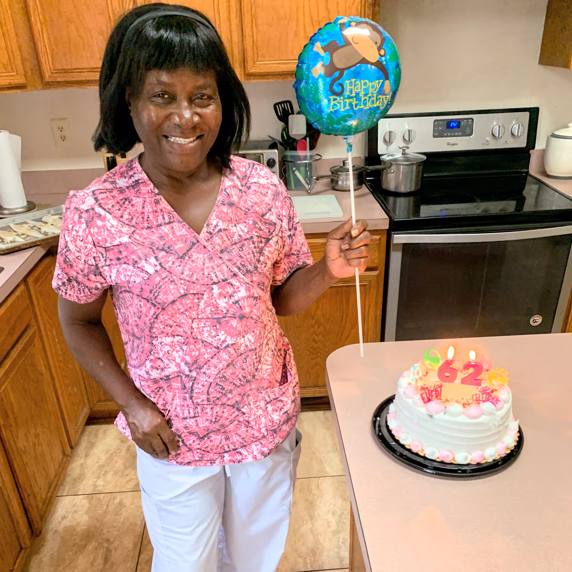 A smiling woman in a pink patterned scrub top and white pants stands in a kitchen holding a blue balloon with a monkey and the words 'Happy Birthday!'. On the counter in front of her is a birthday cake with lit candles showing the number 62.