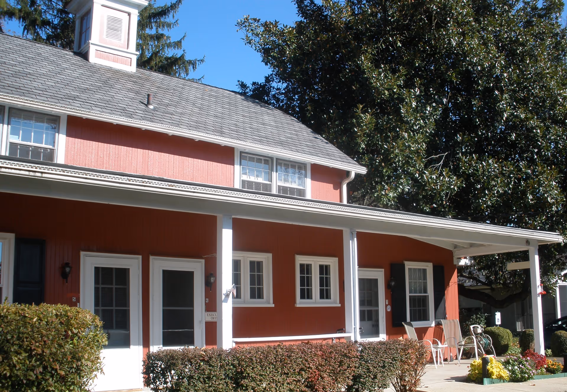 Exterior view of a red building with white trim at Friends Village, Paxson Campus. The building has a covered porch with white columns, several windows, and doors. There are bushes and flowers planted along the front, and outdoor chairs are placed on the porch. A large tree is visible in the background under a clear blue sky.