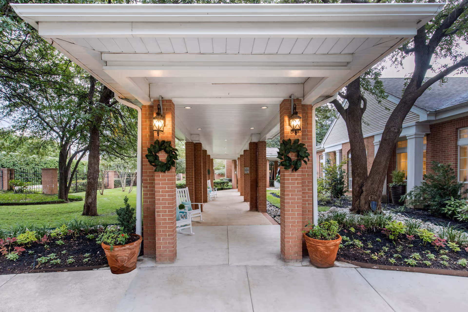 Covered walkway with brick columns decorated with green wreaths, white rocking chairs along the path, and landscaped garden beds with plants and trees on either side.